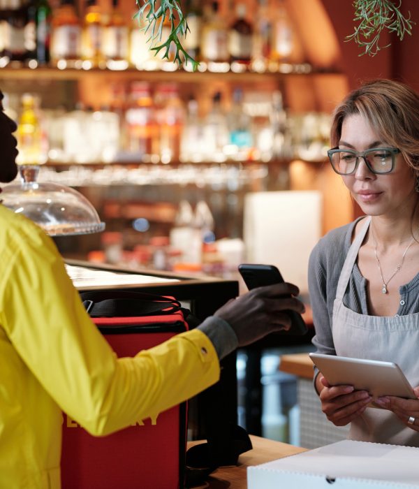 Young waitress in apron looking at list of orders on screen of smartphone held by African American courier in helmet and yellow jacket